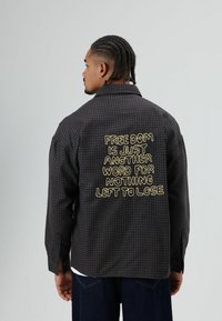 Zwart geruit overhemd met gele geborduurde tekst op de achterkant. Ontwerp met knoopsluiting, lange mouwen en zijsplitsingen. Gemaakt van katoen.