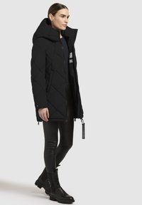 Must musträrgiga puffer mantel kõrge kraega, esi tõmblukk ja küljetaskud. Kandas mustade nahkleggingite ja pahkluudeni saapatega.