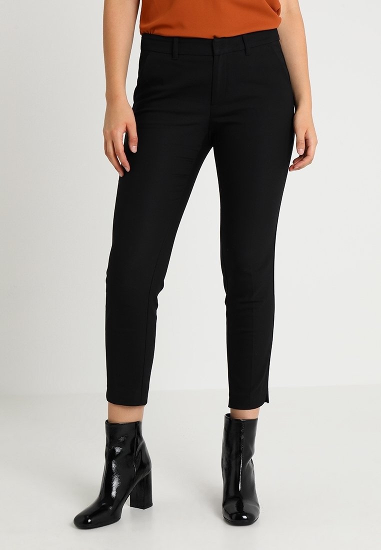 Femme portant un pantalon noir ajusté aux chevilles, des bottines noires brillantes et un haut orange sur un fond uni.