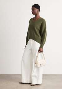 Olivgrön stickad tröja med V-hals, i kombination med vida vita byxor och en krämfärgad handväska. Modellen bär hängande örhängen och låga skor.