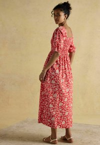 Rotes maxikleid mit Blumenmuster, Puffärmeln, quadratischem Rücken und strukturiertem Stoff. Kombiniert mit schlichten flachen Sandalen, vor einem neutralen Hintergrund.