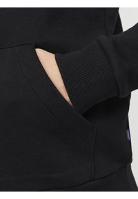 Main insérée dans la poche avant d’un sweat-shirt noir avec poignets côtelés et étiquette de marque visible sur la couture latérale.