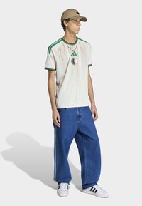 Crèmekleurig voetbalshirt met groene en rode accenten, voorzien van een rond logo. Gecombineerd met losse blauwe spijkerbroeken en witte sneakers met zwarte strepen.