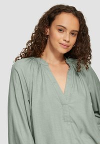 Mintgrünes Hemd mit V-Ausschnitt, langen Ärmeln und gerafften Details an den Schultern; aus einem leichten, strukturierten Stoff gefertigt.