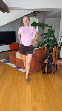 Jeune femme debout, les mains sur les hanches, dans un salon avec un sol en bois, un canapé marron, une plante verte et un rameur à proximité.