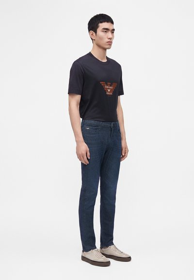 Άνδρας στέκεται φορώντας μαύρο μπλουζάκι Emporio Armani, μπλε τζιν και μπεζ αθλητικά παπούτσια σε απλό φωτεινό φόντο.