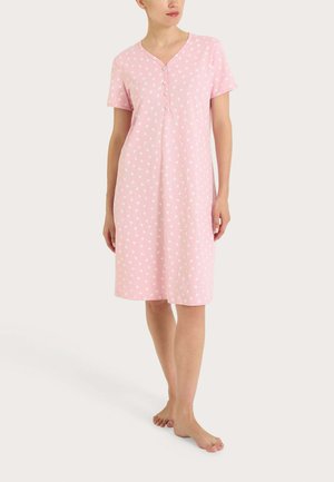 Donna in piedi a piedi nudi che indossa una camicia da notte rosa a maniche corte, lunga fino al ginocchio, con piccoli motivi floreali bianchi e bottoni frontali.