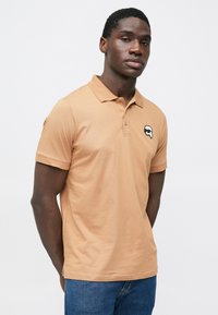 Világosbarna pamut galléros póló rövid ujjakkal és egy rajzfilmfigura logóval a bal mellrészen. Kék farmerrel viselve.