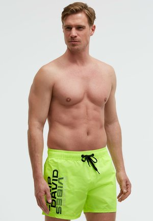 Shirtless man wearing bright yellow-green swim shorts with black drawstring and black text "DAVID SEWING" on left leg standing against plain background.