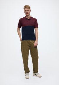 Polo shirt in burgundy and navy, short sleeves, with a collar and a small logo. Paired with olive green trousers and light-colored sneakers.