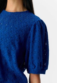 Blauwe kanten top met een textuurrijk diamantenpatroon, korte puffmouwen en een ronde halslijn, die de ingewikkelde stofdetails benadrukt.