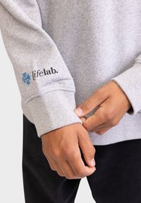 Felpa grigia con polsini a coste, con una grafica blu e la scritta "lifelab." sulla manica. Il tessuto appare morbido e strutturato.