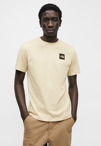 Ανοιχτό μπεζ βαμβακερό t-shirt με κοντά μανίκια και στρογγυλή λαιμόκοψη, με μικρό καφέ λογότυπο στην αριστερή πλευρά του στήθους.