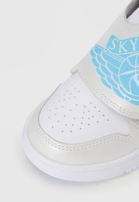 Baltas un sudraba kedas ar perforētu pirkstu daļu, zilu spārnu logotipu un Velcro siksniņu. Gluda ādas līdzīga materiāla, noapaļota forma.