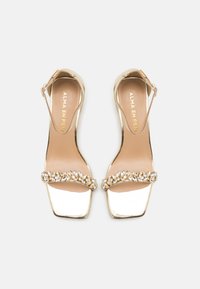 Sandalias de tacón alto metálicas doradas con punta cuadrada, que cuentan con una tira decorativa de piedras de strass y un cierre de hebilla en el tobillo.