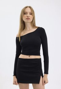Schwarzes langärmeliges Oberteil mit einem freiliegenden Schulter, gerafftem Design an der Taille. Kombiniert mit einem eng anliegenden schwarzen Minirock mit seitlicher Raffung.
