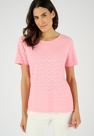 Vrouw draagt een roze korte mouwen eyelet top en witte broek, staat tegen een neutrale achtergrond, glimlacht licht naar de camera.