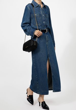Vrouw draagt een lange denim overhemdjurk met een voorste split, een zwarte riem, een zwarte schoudertas met gouden ketting en zwarte hoge hakken mules.