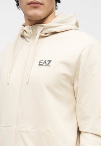 Bărbat purtând hanorac crem cu fermoar, glugă și șnururi, cu logo negru EA7 Emporio Armani pe partea stângă a pieptului.