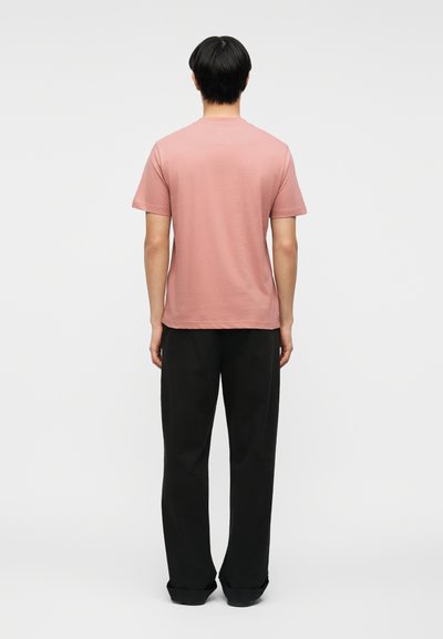 Άνδρας που φοράει ένα ροζ t-shirt με κοντά μανίκια και μαύρο παντελόνι με φαρδιά μπατζάκια, στέκεται πλάτη σε απλό φόντο.