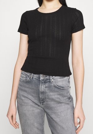 Kvinde iført en tætsiddende sort kortærmet striktop med subtile lodrette mønstre kombineret med højtaljede, falmede grå jeans.