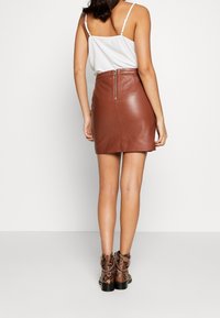 Brauner Minirock aus Leder mit glatter Oberfläche und Reißverschluss hinten, kombiniert mit einem weißen Trägertop und knöchelhohen Stiefeln mit Schlangenprint.