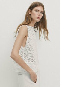 Jeune femme aux longs cheveux, portant un top blanc en dentelle sans manches et un pantalon blanc, debout devant un fond clair uni.