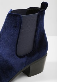 Botines de terciopelo azul marino con paneles laterales elásticos y tacón negro en bloque. Cuentan con una textura suave y líneas de diseño limpias.