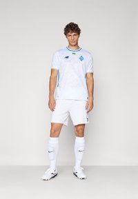 Hvit fotballtrøye med lyseblå detaljer, med et geometrisk mønster, korte ermer og laglogo. Kombinert med hvite shorts og sokker.