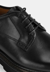 Chaussure habillée en cuir noir avec une finition lisse, un système de laçage et des coutures détaillées le long des coutures et de la semelle.