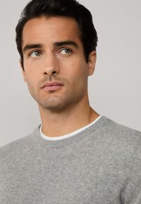 Szary dziany sweter z okrągłym dekoltem, teksturowanym materiałem i białym wewnętrznym kołnierzykiem. Modelka ma gładką skórę i ciemne włosy.