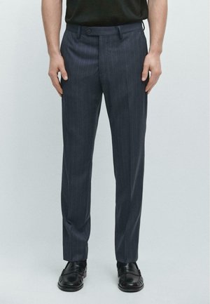 Mann trägt marineblaue Nadelstreifen-Hose und schwarze Leder-Loafer und steht vor einem schlichten hellen Hintergrund.