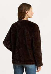 Barna műszőrméből készült dzseki, puha tapintású, laza fazonú, kerek nyakú, széles ujjak és a deréknál végződő alj, kék farmernadrággal párosítva.