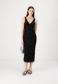 Μαύρο midi φόρεμα με ανθιστή υφή, V-ανοιχτό λαιμό και λεπτές τιράντες, συνδυασμένο με μαύρες σανδάλες και ασημένια τσάντα. Λείο φινίρισμα υφάσματος.