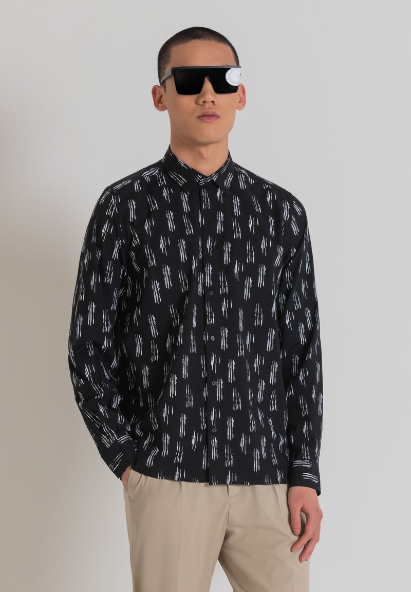 Camisa negra de botones con rayas verticales blancas, de mangas largas y con un cuello clásico. Lleva unas gafas de sol negras de gran tamaño.
