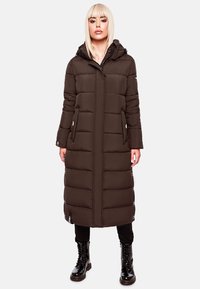 Bruine pufferjas met een capuchon, met horizontaal gestikte secties, zijzakken en drukknoopdetails. Draagt over zwarte broek en laarzen.