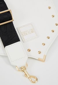 Detal białej skórzanej torby z złotymi ćwiekami, złotymi ozdobami na rogach oraz czarnym regulowanym paskiem z złotą klamrą i zapięciem, metka Versace Jeans Couture.