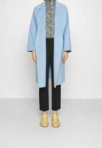 Světle modrý kabát s širokým límcem, černý a bílý vzorovaný top, černé kalhoty a žluté mokasíny s texturovaným designem.