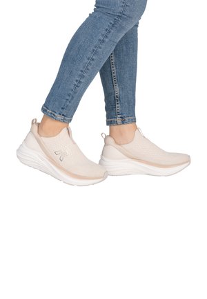 Personne portant des baskets beige clair à enfiler avec des semelles blanches épaisses et un jean bleu sur fond blanc.
