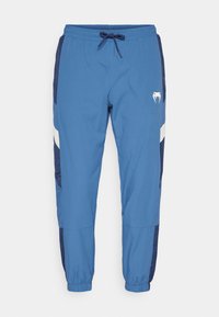 Blauwe joggingsbroek met een trekkoord in de taille, voorzien van marineblauwe zijpanelen, witte strepen en een logo nabij de heup. Elastische boorden bij de enkels.