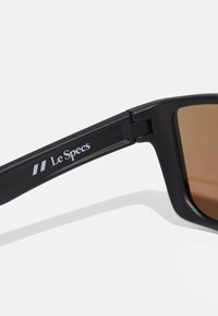 Mustat aurinkolasit matalla pinnalla, ruskeat sävytetyt linssit ja hopeinen "Le Specs" -logo varressa. Kulmikas kehysmuotoilu.