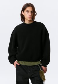 Pull noir oversize avec col et poignets côtelés, superposé à une chemise vert foncé. Comprend un porte-clés avec un accent en fourrure.