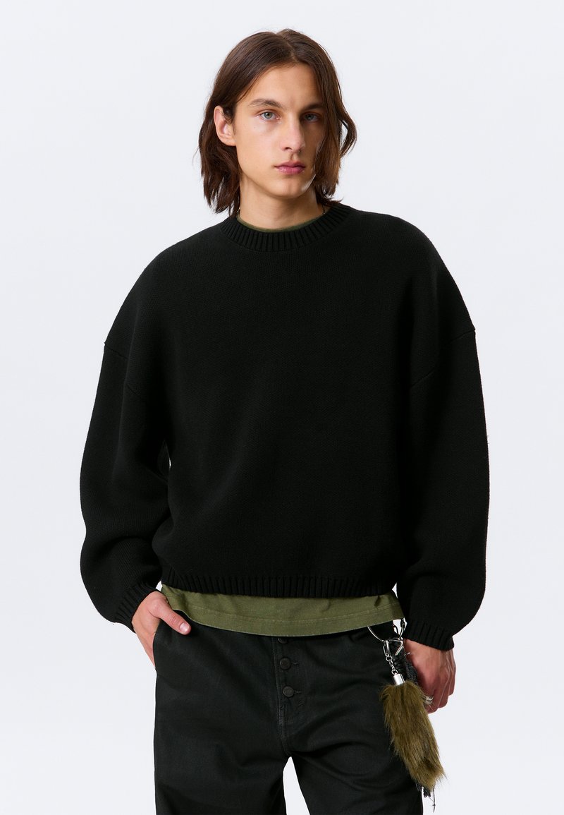 Pull noir oversize avec col et poignets côtelés, superposé à une chemise vert foncé. Comprend un porte-clés avec un accent en fourrure.