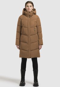 Pitkä ruskea untuvaparkatakki, jossa on huppu, vetoketjukiinnitys ja nappikiinnitys, palotettu muotoilu ja sivutaskut, käytetty mustien leggingsien ja pitkien saappaiden päällä.