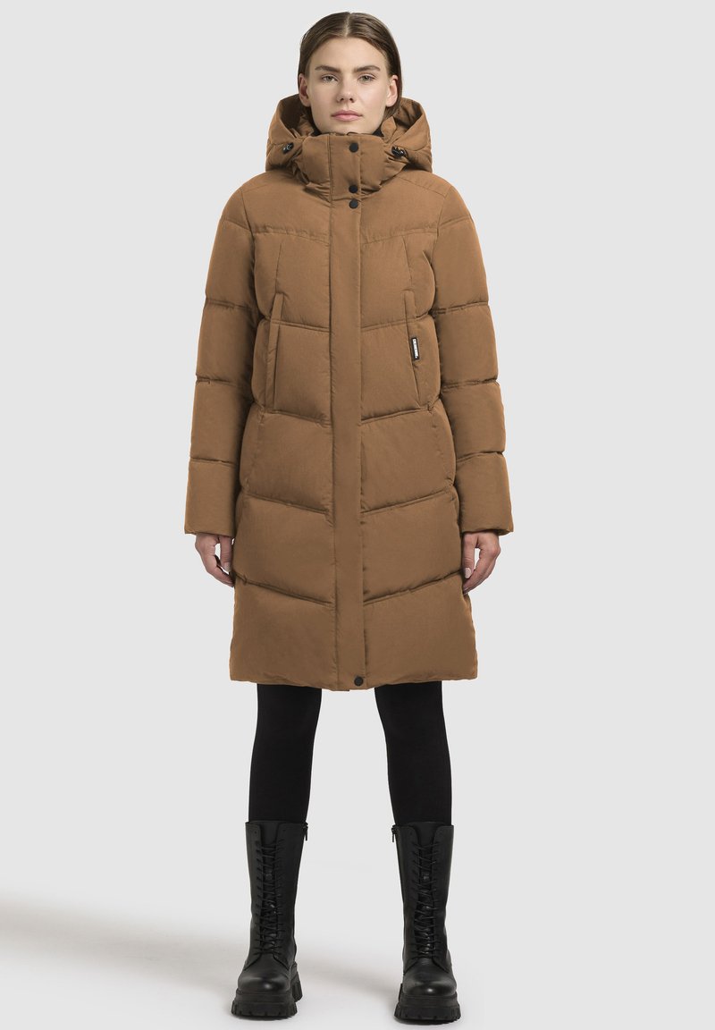 Pitkä ruskea untuvaparkatakki, jossa on huppu, vetoketjukiinnitys ja nappikiinnitys, palotettu muotoilu ja sivutaskut, käytetty mustien leggingsien ja pitkien saappaiden päällä.