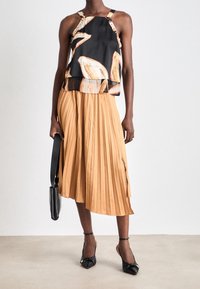 Mujer con un top halter abstracto en negro y beige, falda midi plisada color beige, tacones negros con correa en el tobillo, sosteniendo un bolso negro frente a un fondo blanco.