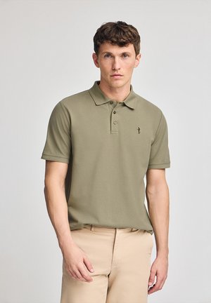 Polo color verde oliva con cuello, dos botones y un logotipo bordado en el lado izquierdo del pecho. Mangas cortas, textura suave, combinado con pantalones beige.