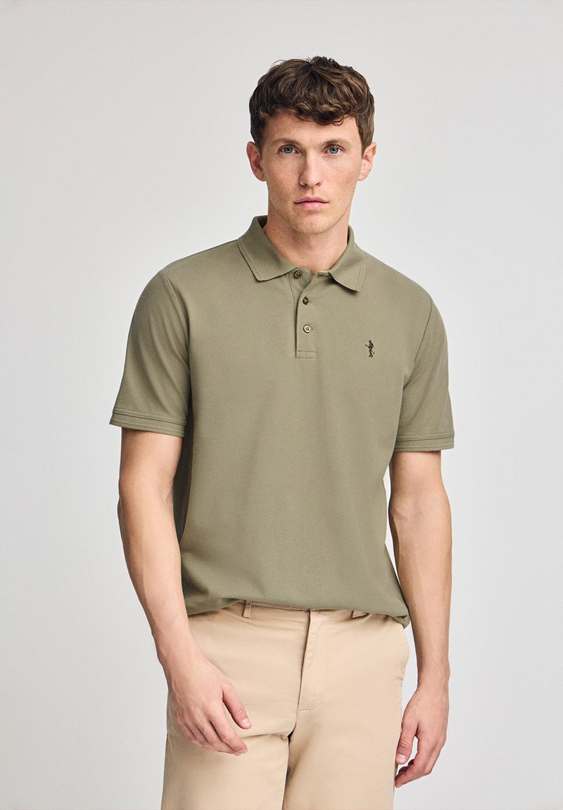 Olivgrön polotröja med krage, två knappar och en broderad logotyp på vänster bröst. Kortärmad, mjuk textur, tillsammans med beige byxor.