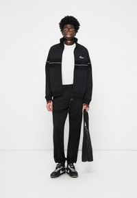 Sort tracksuit jakke med hvide striber, parret med sorte bukser og sneakers. Holder en sort taske. Ensfarvede farver, afslappet pasform og sporty design.