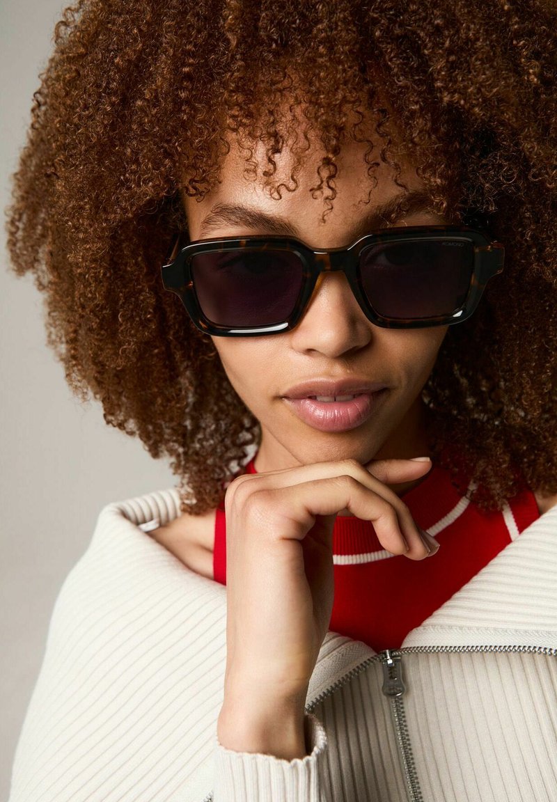 Bruine schildpadzonnebril met donkere lenzen, hoekige vorm en minimale metalen accenten; gedragen met een crèmekleurige geribbelde jas en een rode top.